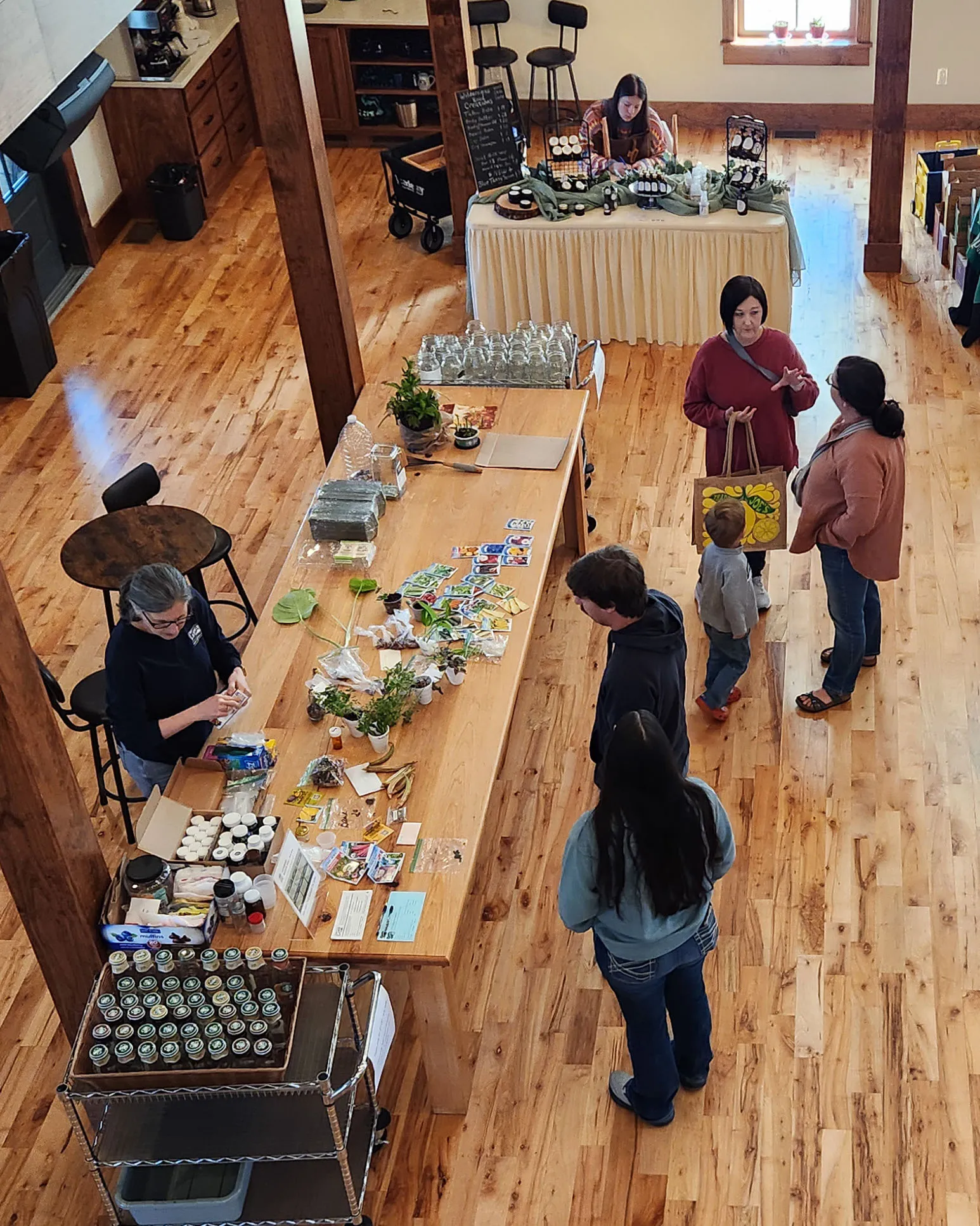 Seen from the balcony, visitors browse a long wooden table of plants and crafts in the hall.