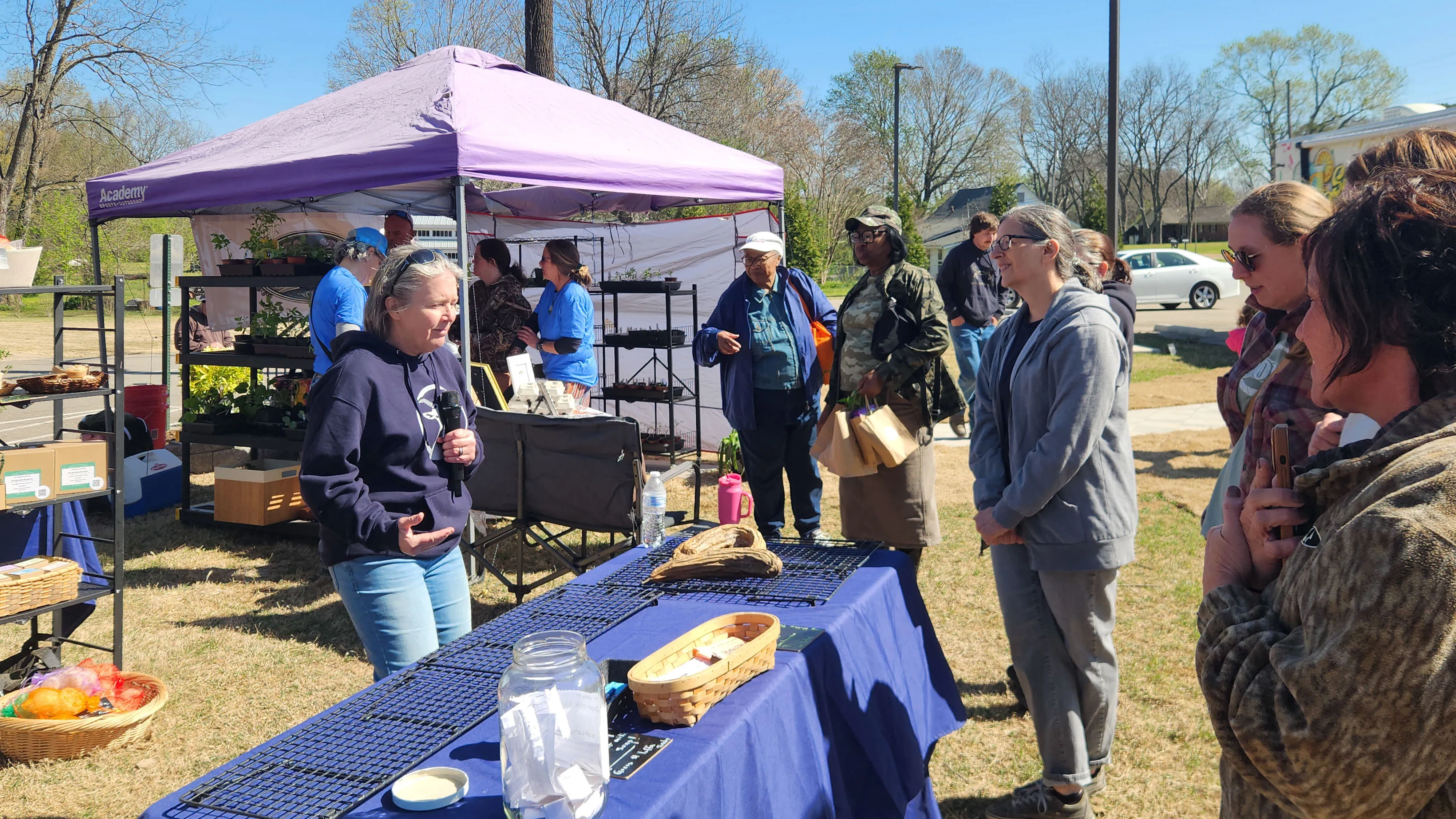 A vendor under a purple tent talks with visitors beside a blue-covered table of handmade goods.