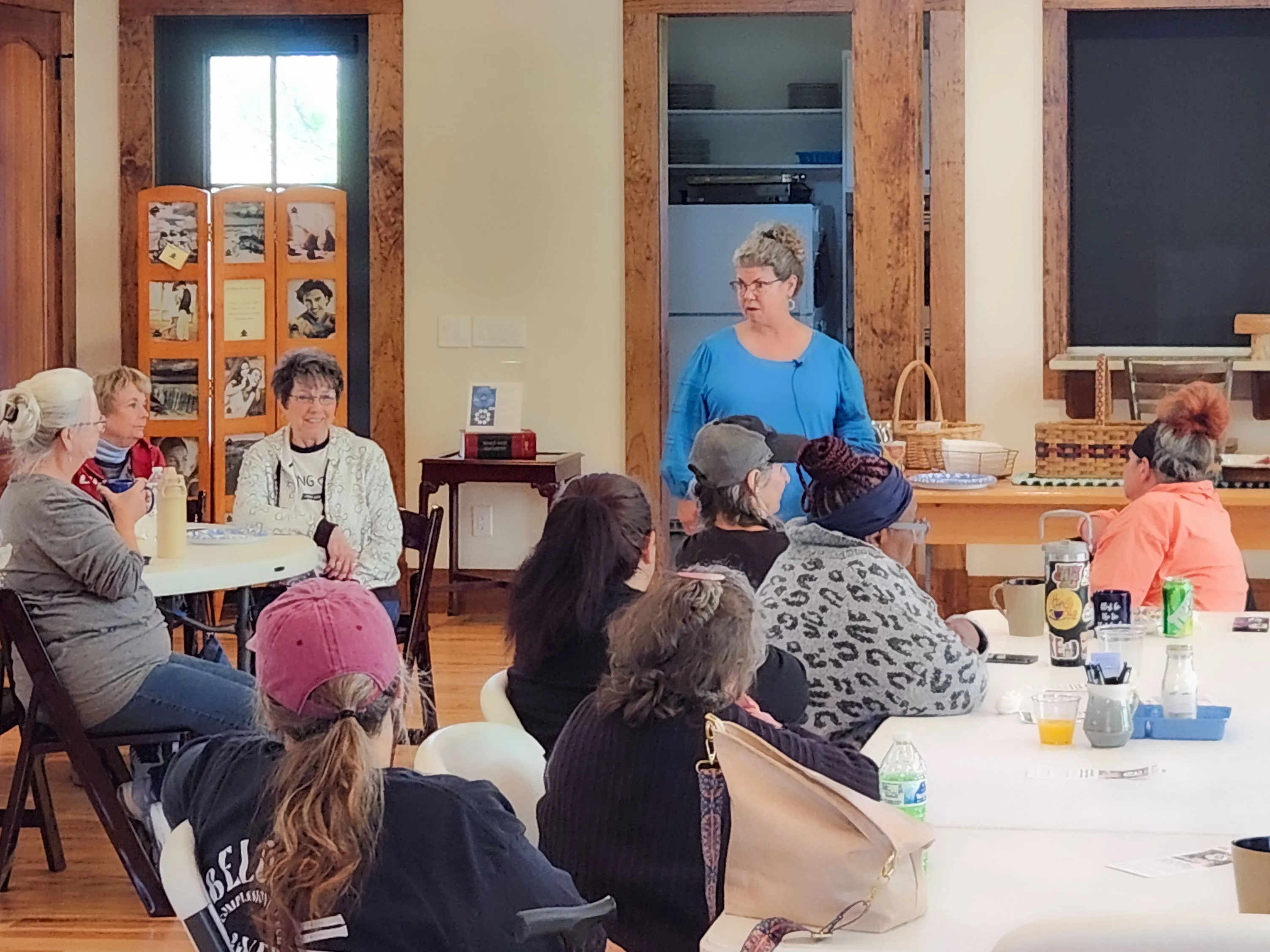 A speaker in a blue top talks with women seated at tables during the Place of Joy morning.