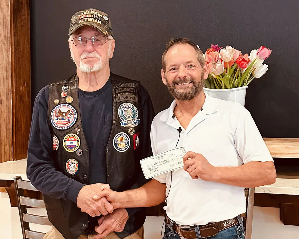 A veteran in a patched vest shakes hands with a man in a white shirt as he hands over a check.