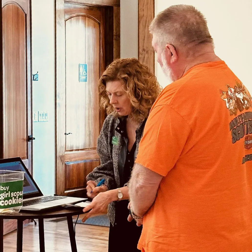 A Girl Scouts representative shows a laptop to a man in an orange shirt next to a sign about cookies.