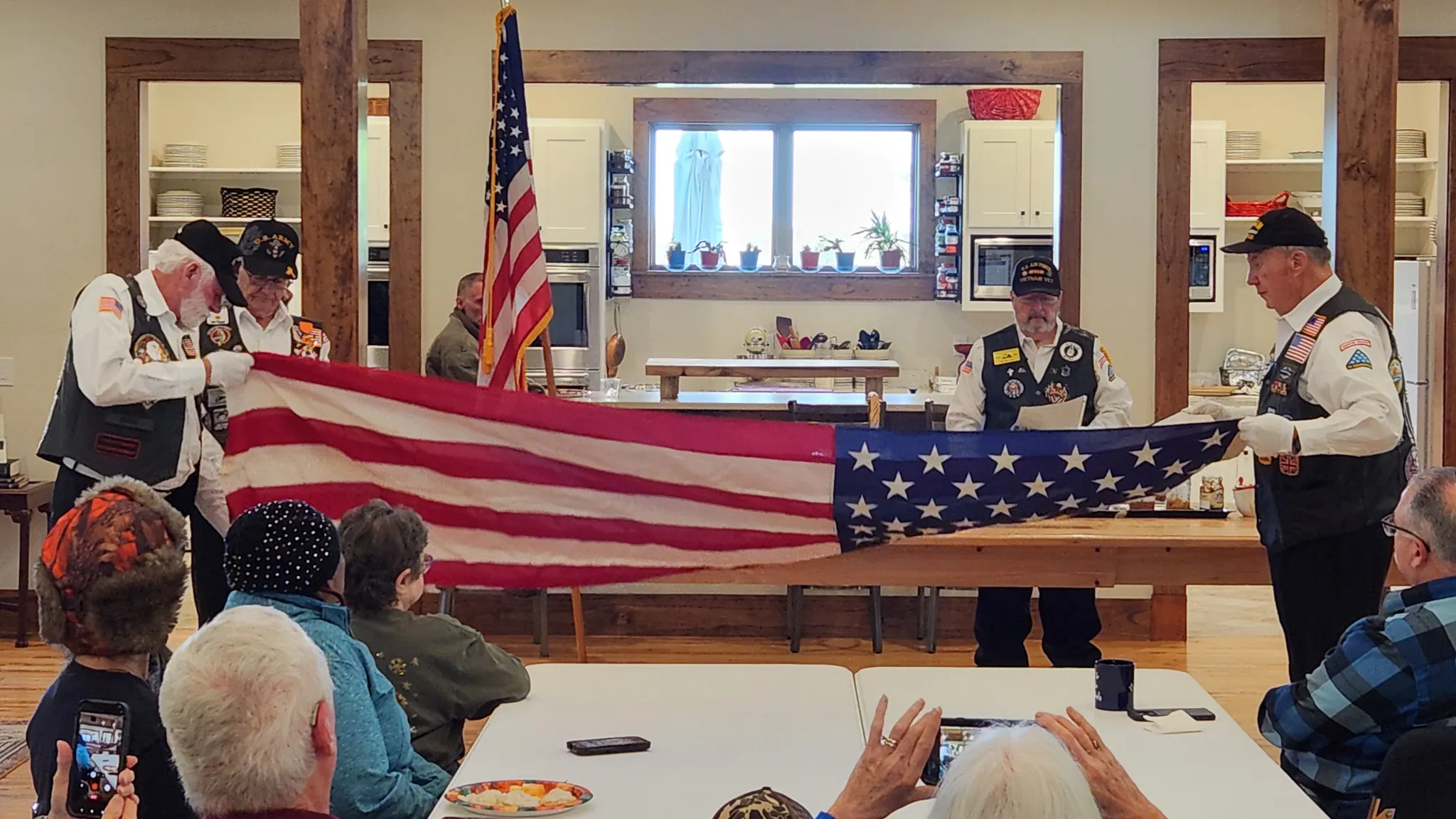 Veterans hold a folded American flag over a table while neighbors watch, with a flag standing in the background.