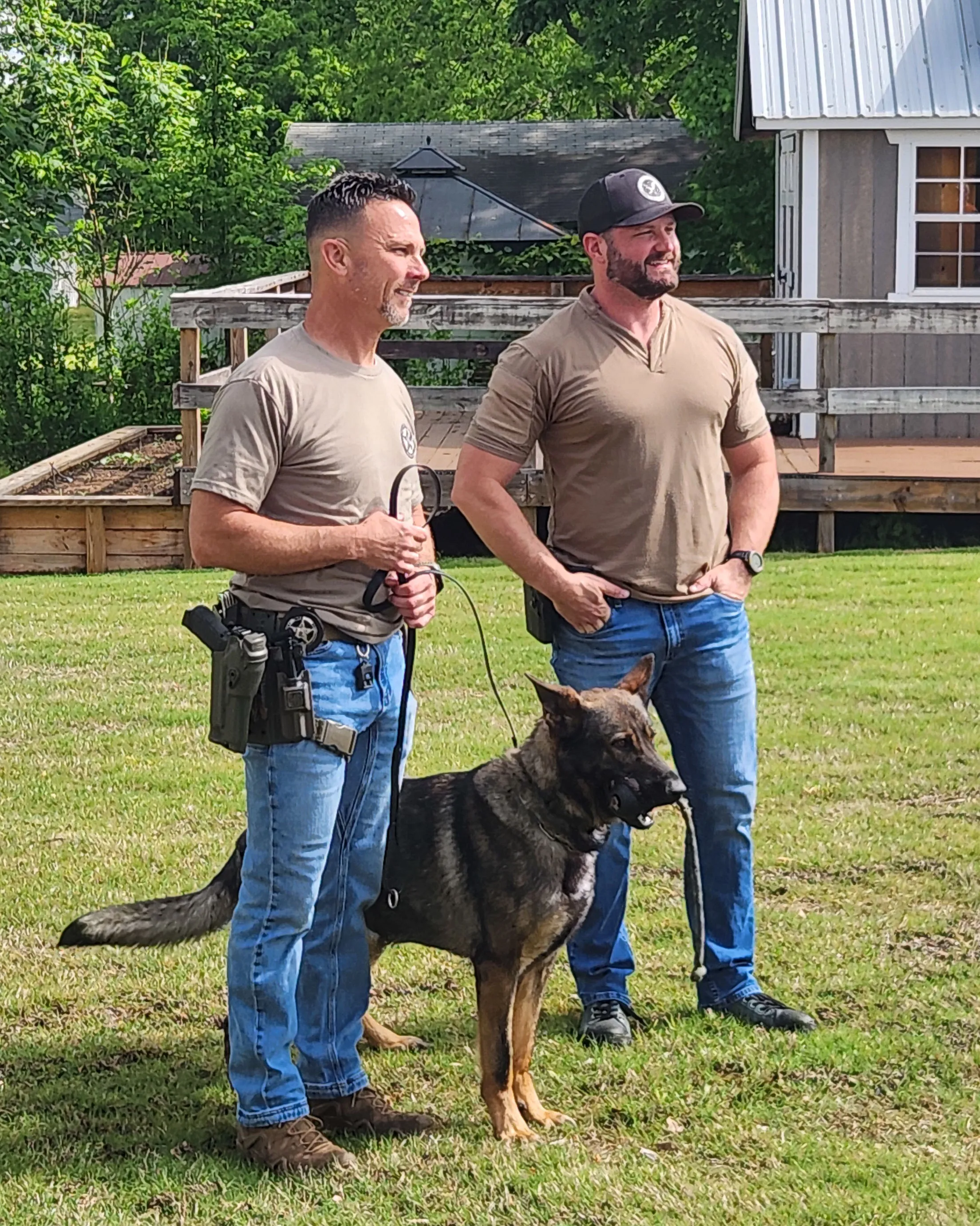 Two officers stand on the lawn with their German shepherd between them.