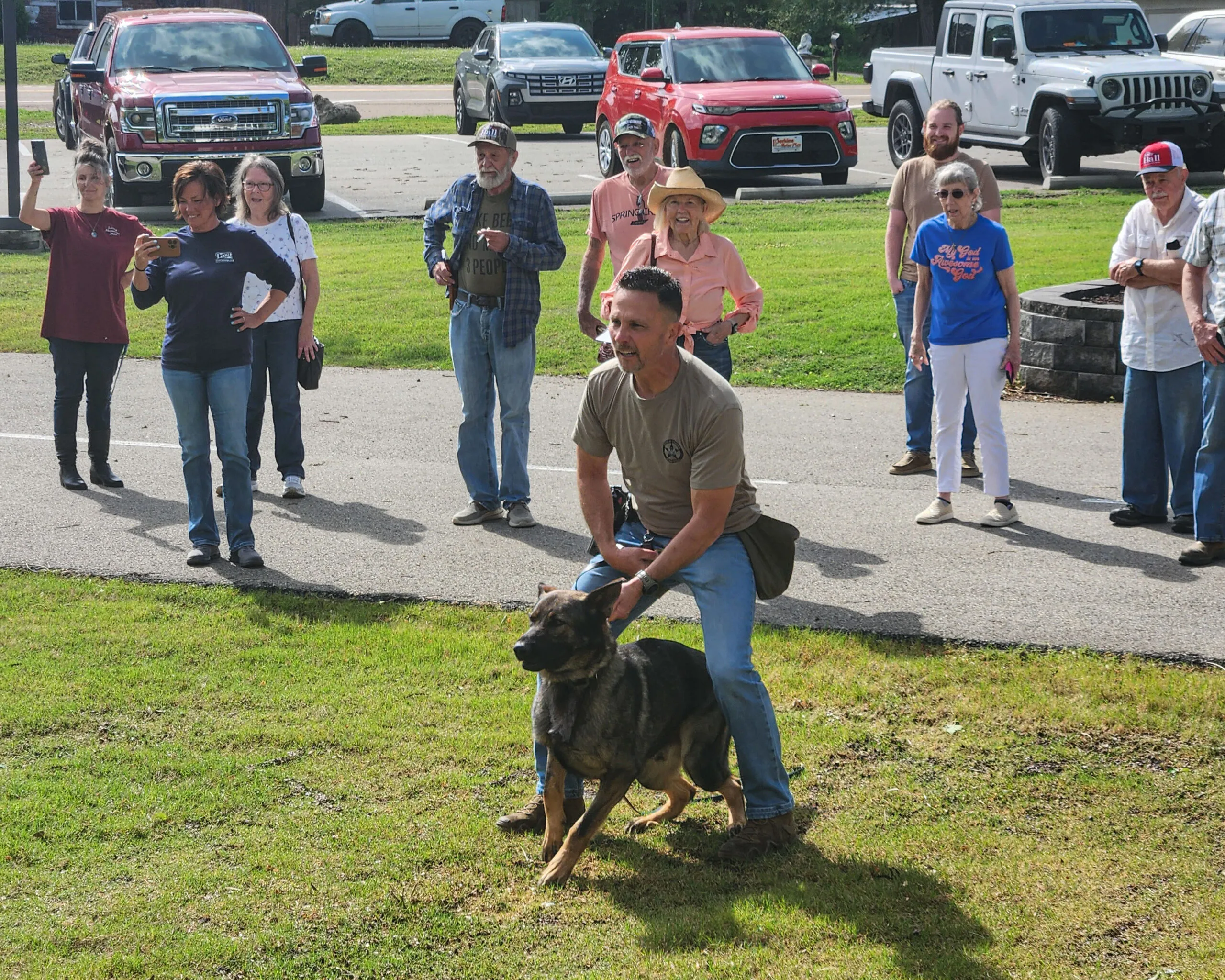 A K9 handler crouches beside his police dog while neighbors watch from the parking area.
