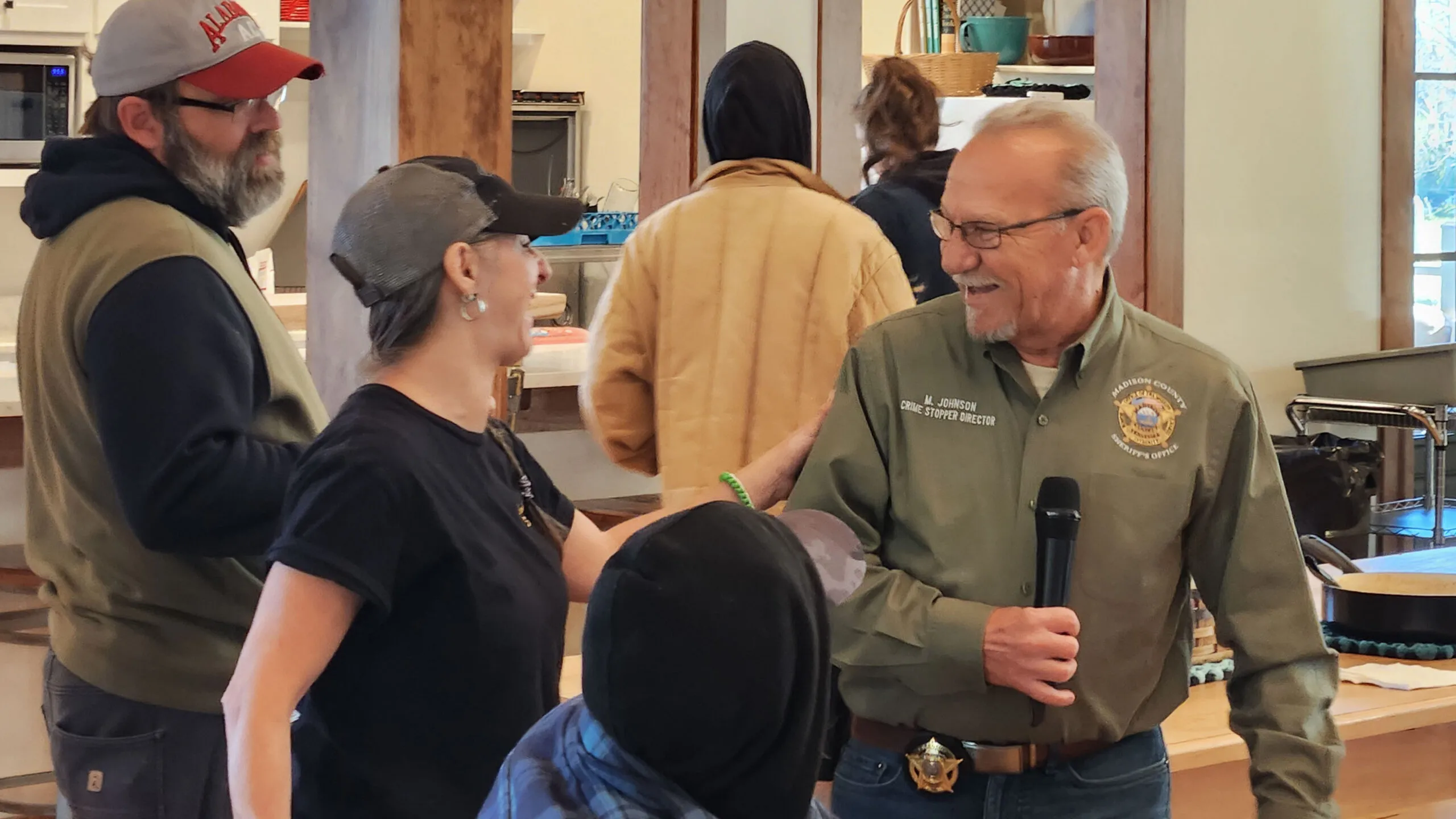 A Crime Stoppers speaker in a tan uniform shirt holds a microphone and smiles as neighbors gather around him.
