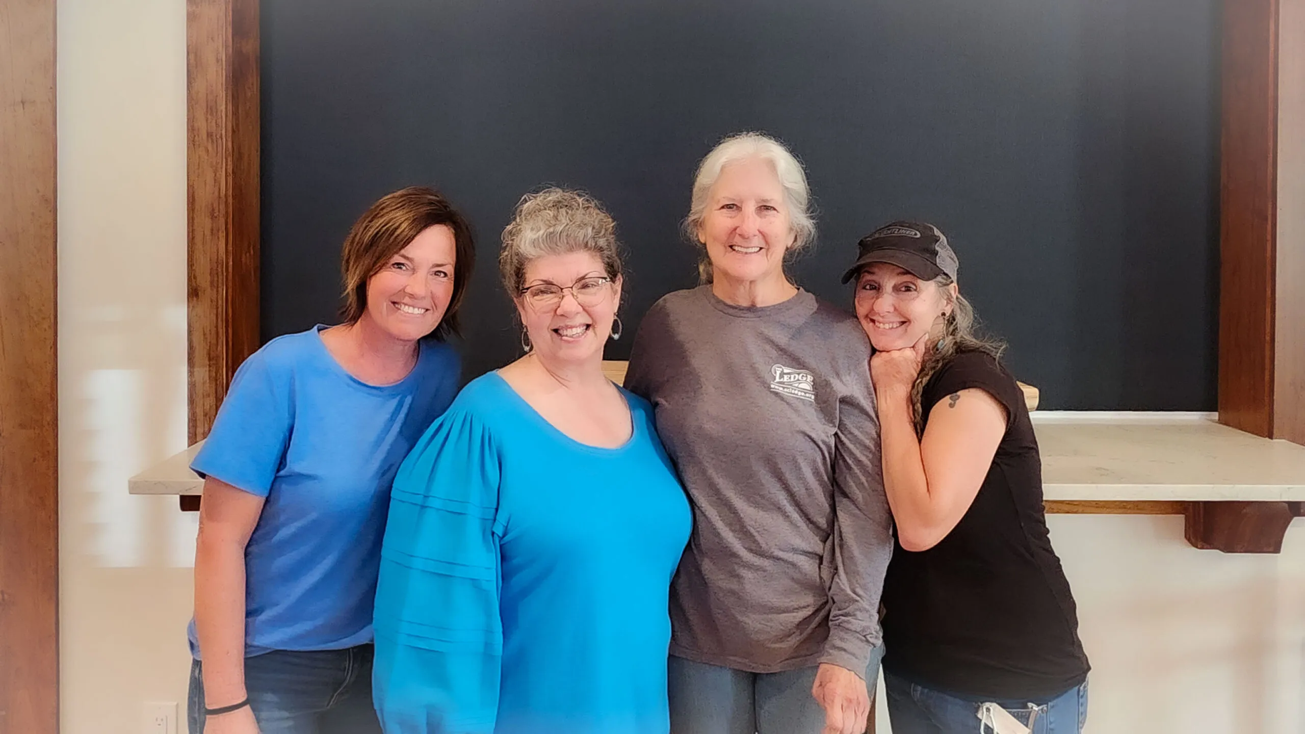 Four women in blue and gray shirts smile together in the kitchen area of The Ledge.