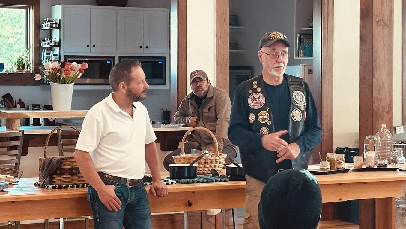 A man in a white shirt and a veteran in a vest covered in patches stand together in the kitchen area during the talk.
