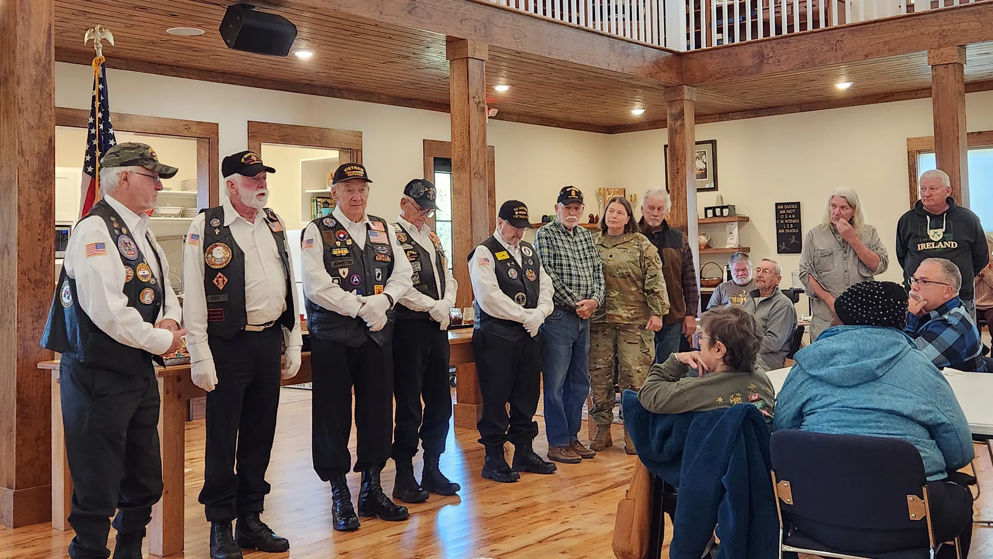 Veterans in black vests and caps stand in a line in the hall during the Veterans Day flag ceremony.