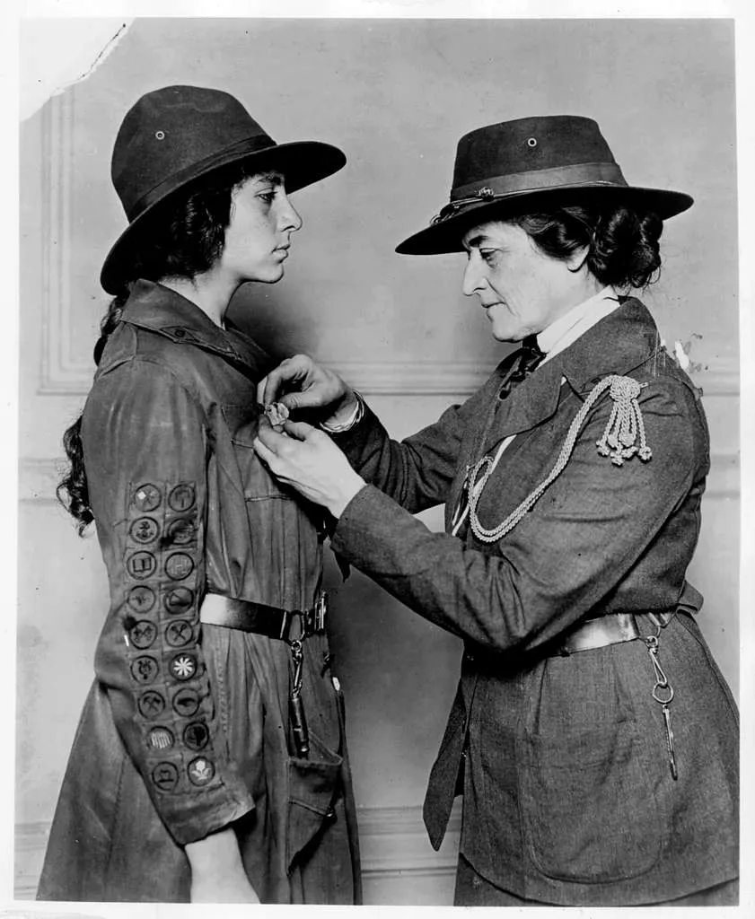 An old black-and-white photo of a Girl Scout leader pinning a badge on a scout’s uniform sleeve.